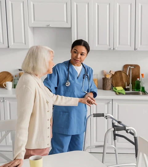 A nurse in blue scrubs, with a stethoscope, assists an elderly woman with a walker in a bright kitchen, conveying care and support.