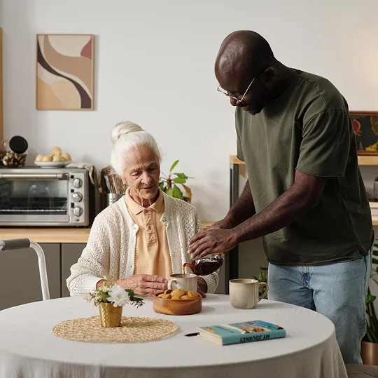 Male caregiver pouring tea for an elderly woman at a kitchen table