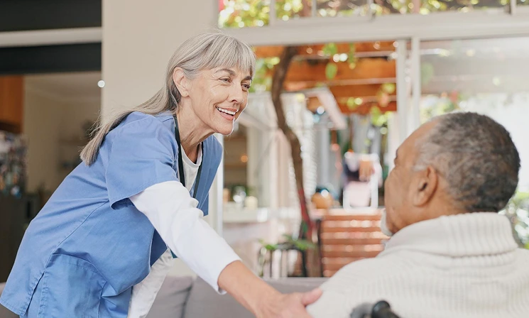 A caregiver in blue scrubs smiles warmly while holding the hand of an elderly man in a wheelchair, creating a friendly and supportive atmosphere.