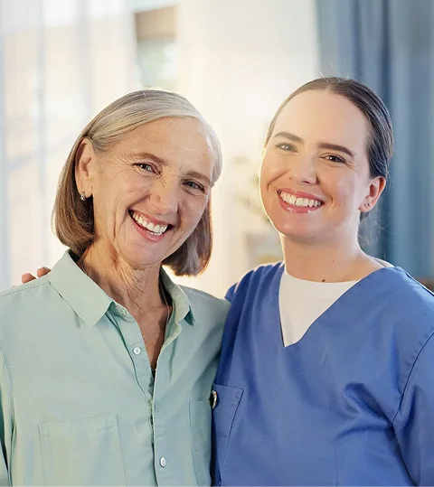 Elderly woman and young nurse in blue scrubs standing together and smiling
