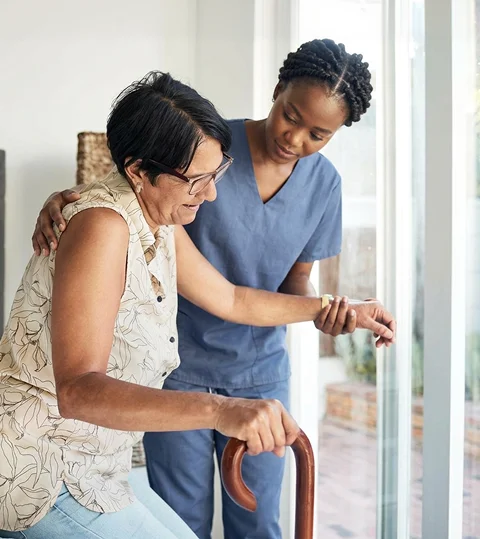 Caregiver in blue scrubs assisting a woman with a cane to stand up