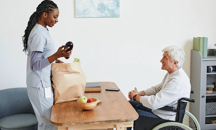 A caregiver in scrubs unpacks groceries at a table while conversing with an elderly man in a wheelchair. The setting is warm and supportive.