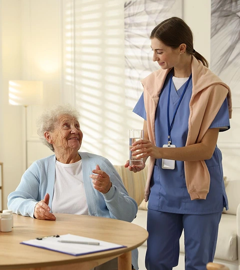 A young nurse in blue scrubs smiles while handing a glass of water to an elderly woman sitting at a table. The setting is bright and warm, conveying care.