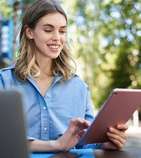 Young woman with blonde hair, wearing a blue shirt, smiling while using a tablet outdoors. Sunlit trees in the background create a serene atmosphere.