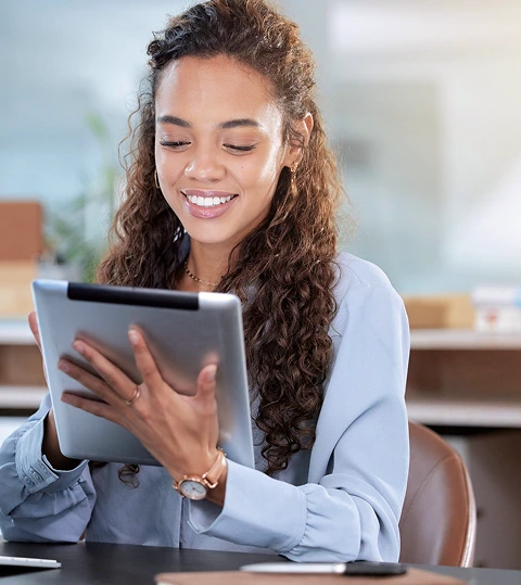 Smiling woman with curly hair in a light blue blouse using a digital tablet. She's seated at a desk in a bright office, conveying a sense of productivity.