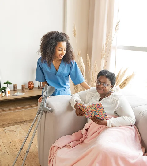 Home caregiver in blue scrubs assisting a patient reading a book on a couch