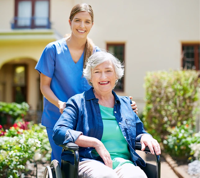 A smiling elderly woman sits in a wheelchair, with a young caregiver in blue scrubs standing behind her.