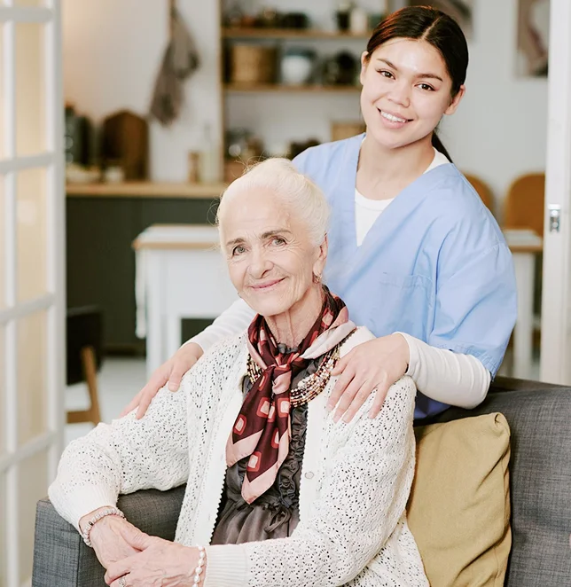 Young caregiver in blue scrubs standing behind a smiling elderly woman seated on a sofa