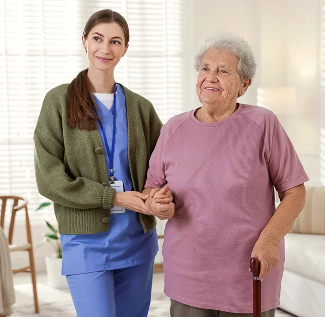 Caregiver holding hands with a smiling elderly woman using a cane indoors