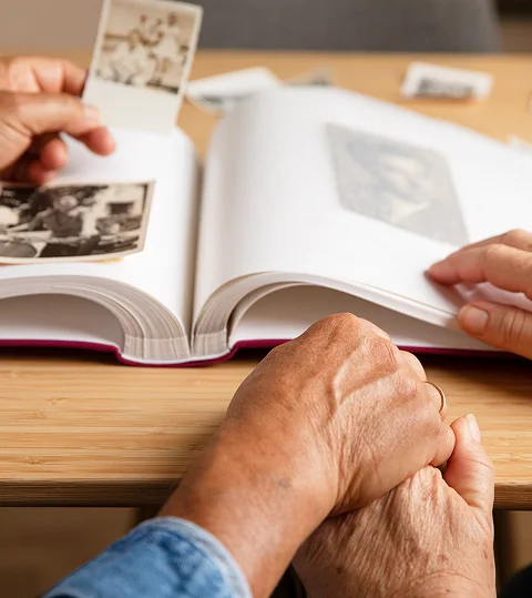 An elderly person's folded hands resting on a table while two people look through an old photo album