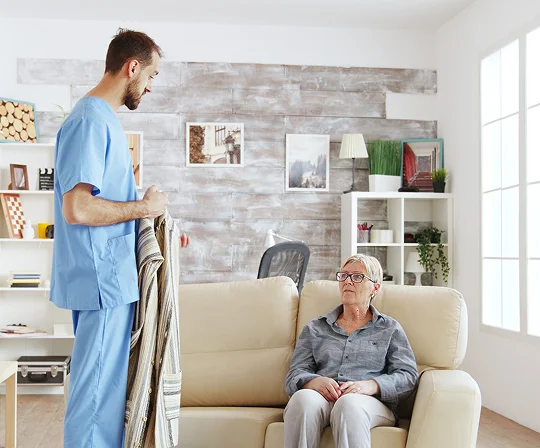 A caregiver bringing a blanket to an elderly woman seated on a sofa