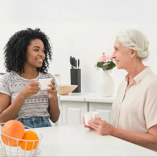 A young woman and an elderly woman sharing coffee and conversation at a kitchen table.