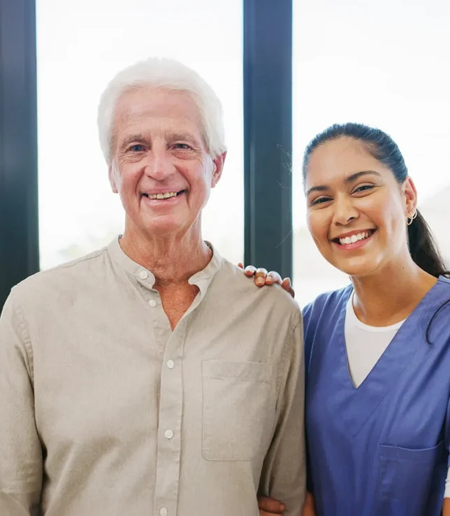 A smiling elderly man and caregiver posing together near a bright window